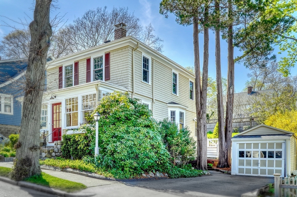Pale yellow two-story house with red shutters, a glassed-in porch, mature trees, and a detached garage.