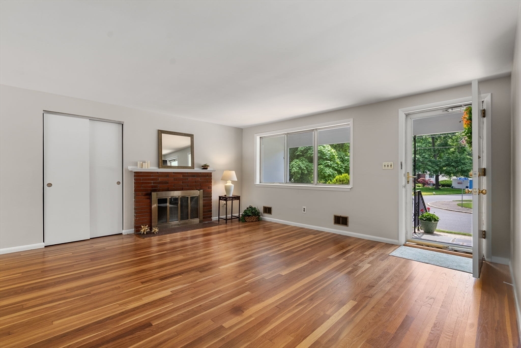 Living room with hardwood floors, brick fireplace, and an open door to a tree-lined street.