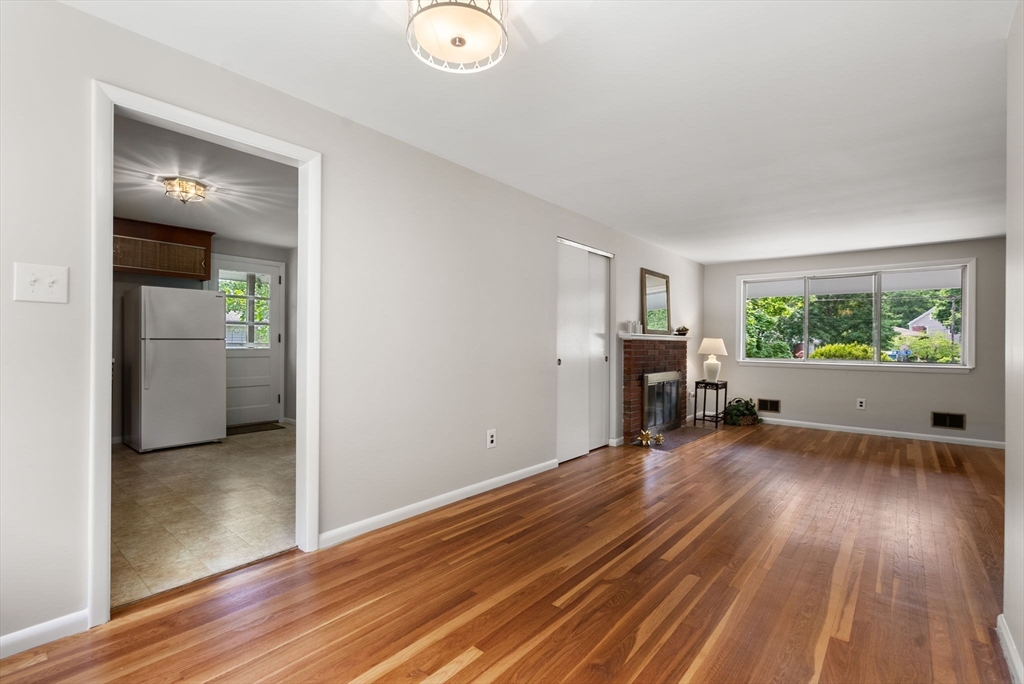 Unfurnished living room with polished hardwood floors, brick fireplace, and wide window overlooking greenery.