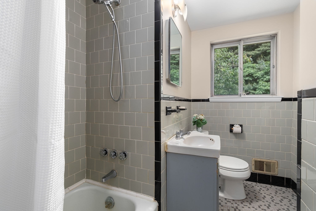 Bathroom with gray tiled tub and shower, gray vanity, patterned floor, and a window overlooking greenery.