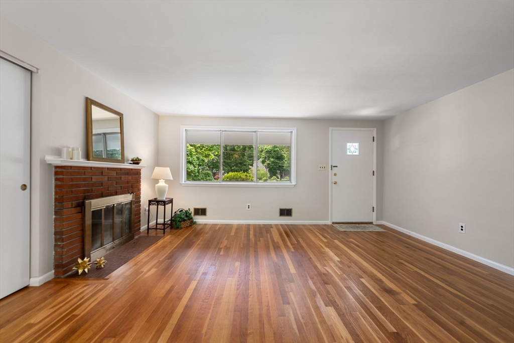 Living room with hardwood floors, a brick fireplace, and a large window overlooking greenery.