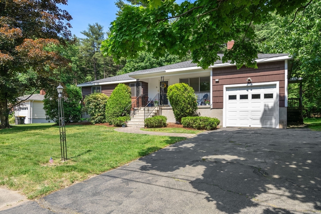Single-story brown home with a white garage door, covered porch, and leafy trees.