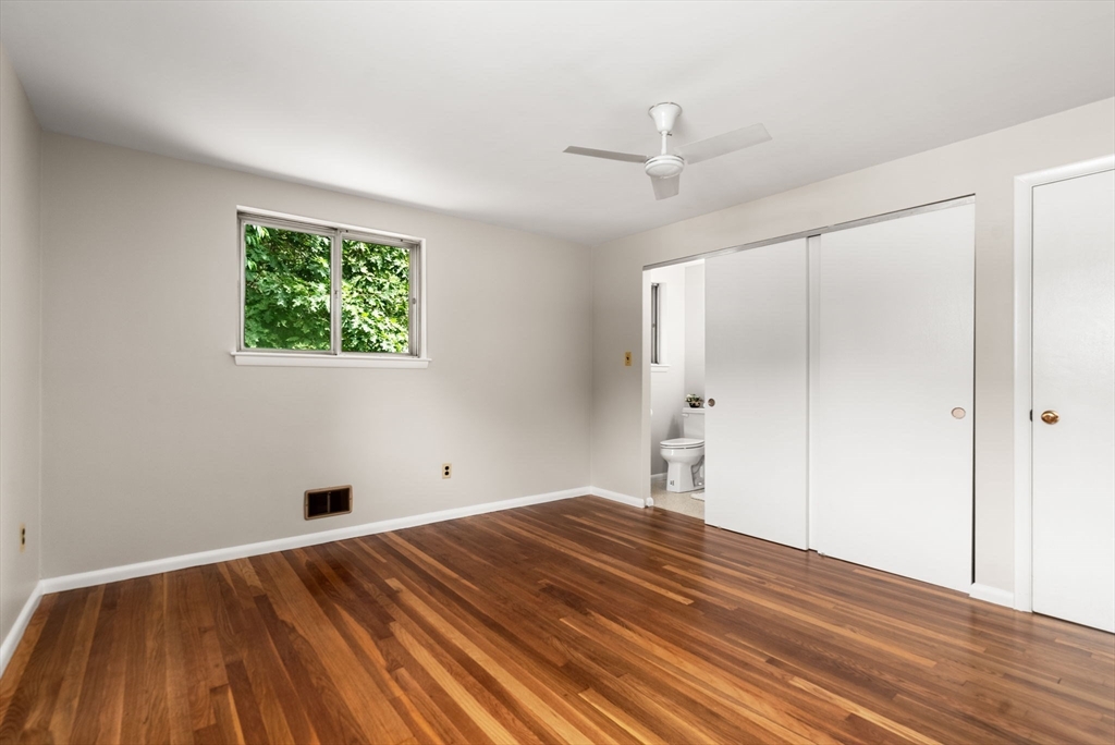 Vacant bedroom with hardwood floors, a ceiling fan, and sliding closet doors beside an open bathroom doorway.