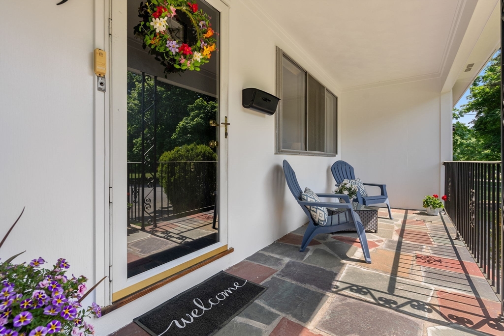 Covered front porch with a colorful wreath on the glass door and two blue Adirondack chairs.