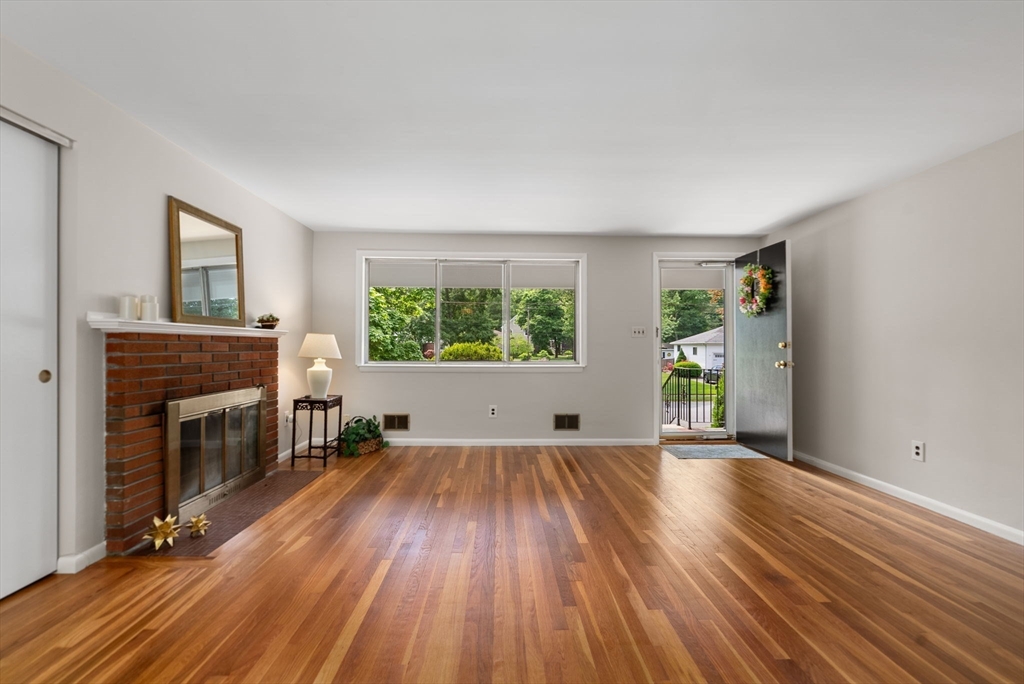 Living room with hardwood floors, brick fireplace, wide windows, and an open front door.