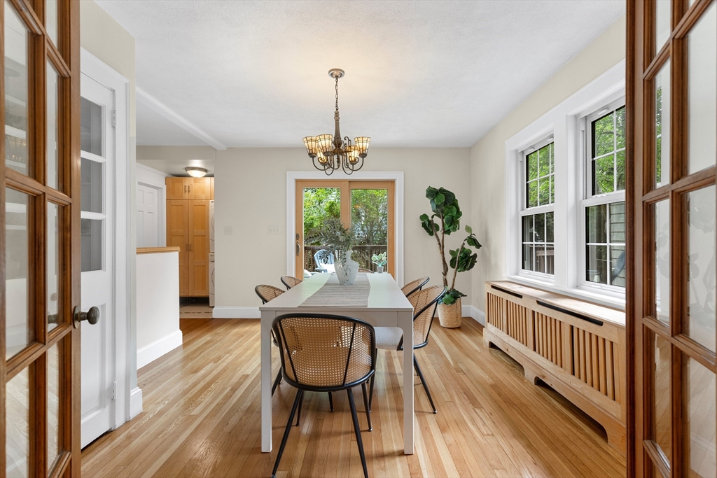 Dining room with a long table, woven chairs, chandelier, and large windows overlooking greenery.