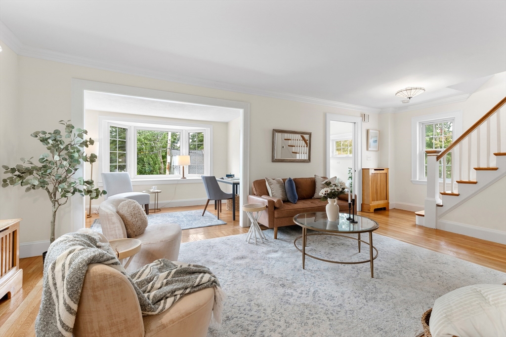 Staged living room with brown sofa, cream armchairs, large rug, and staircase.