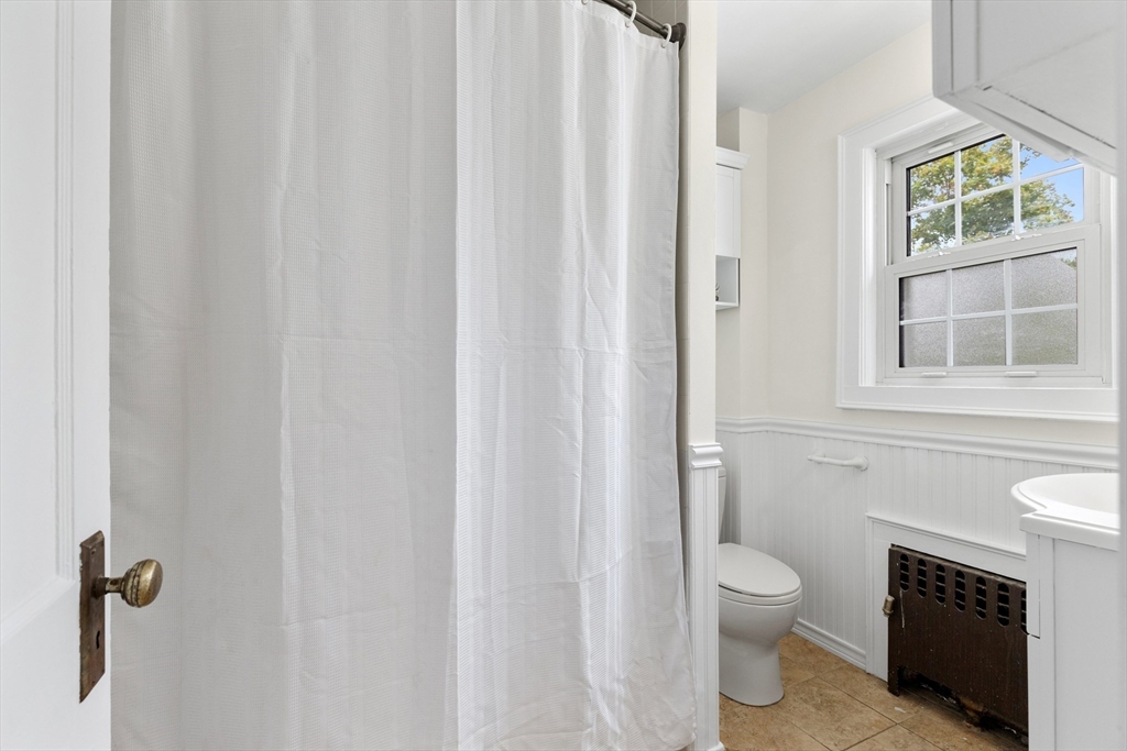 Bathroom with a white shower curtain, toilet, and divided-light window above beadboard wainscoting.