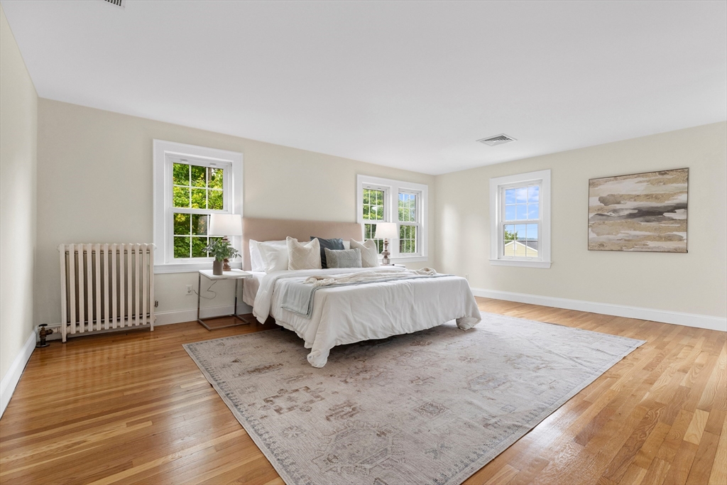 Staged bedroom with a white bed, three windows, hardwood floors, and a pale area rug.