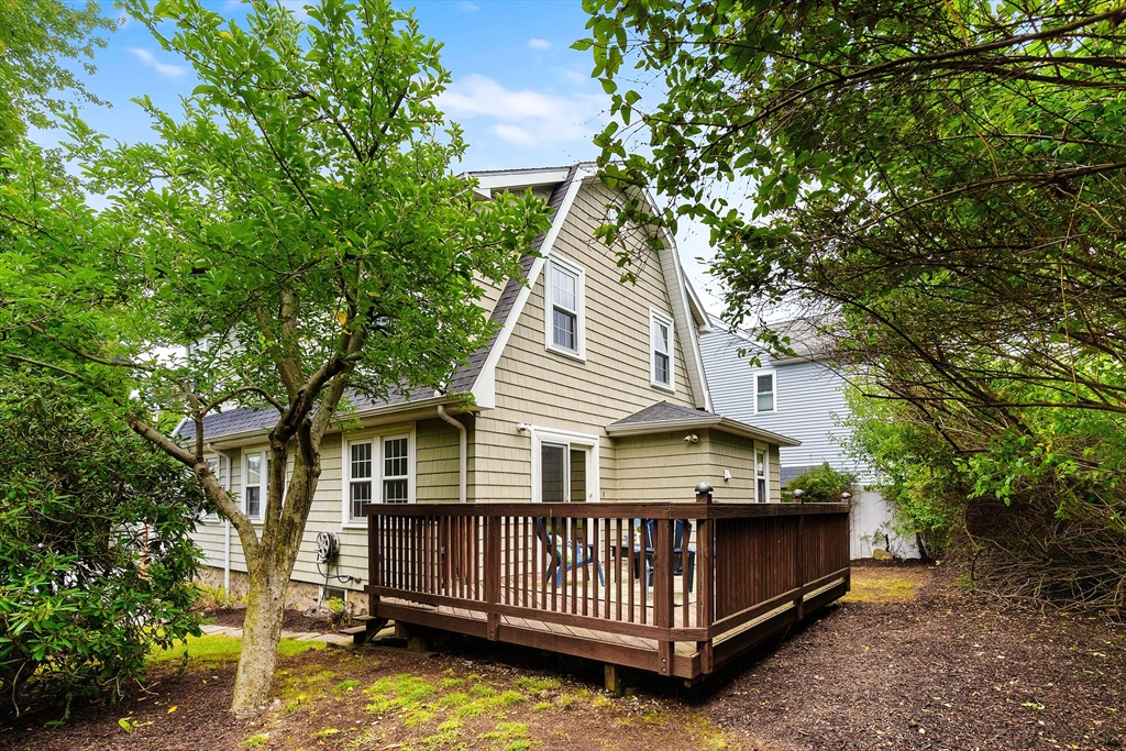 Rear of a beige shingle-sided house with a raised wooden deck framed by leafy trees.