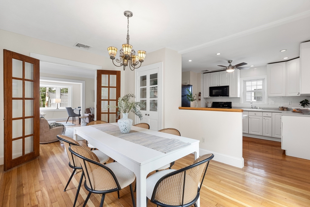 Dining room with a long white table, woven chairs, hardwood floors, and an adjoining white kitchen.