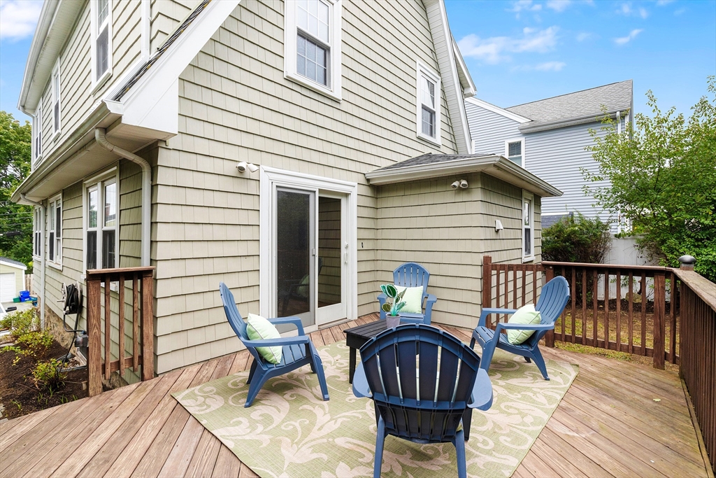 Furnished wooden deck with blue Adirondack chairs and a patterned rug beside a beige-shingled house.