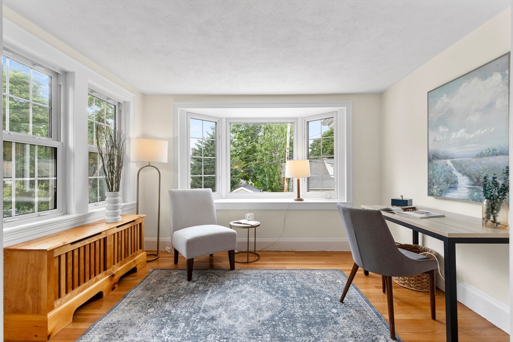 Staged home office with a bay window, gray chairs, blue rug, and wooden cabinet.