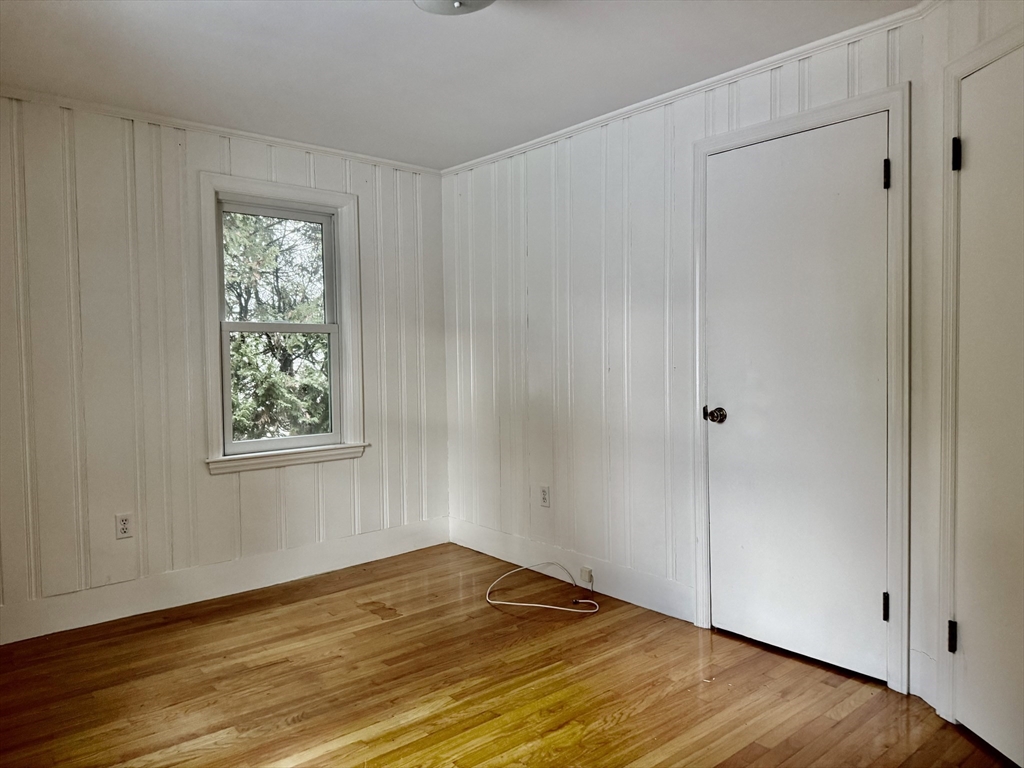 Empty room with white paneled walls, hardwood floors, and a window overlooking trees.