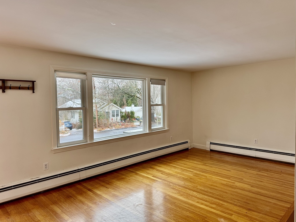 Empty room with hardwood floors, baseboard heating, and wide windows overlooking a residential street.