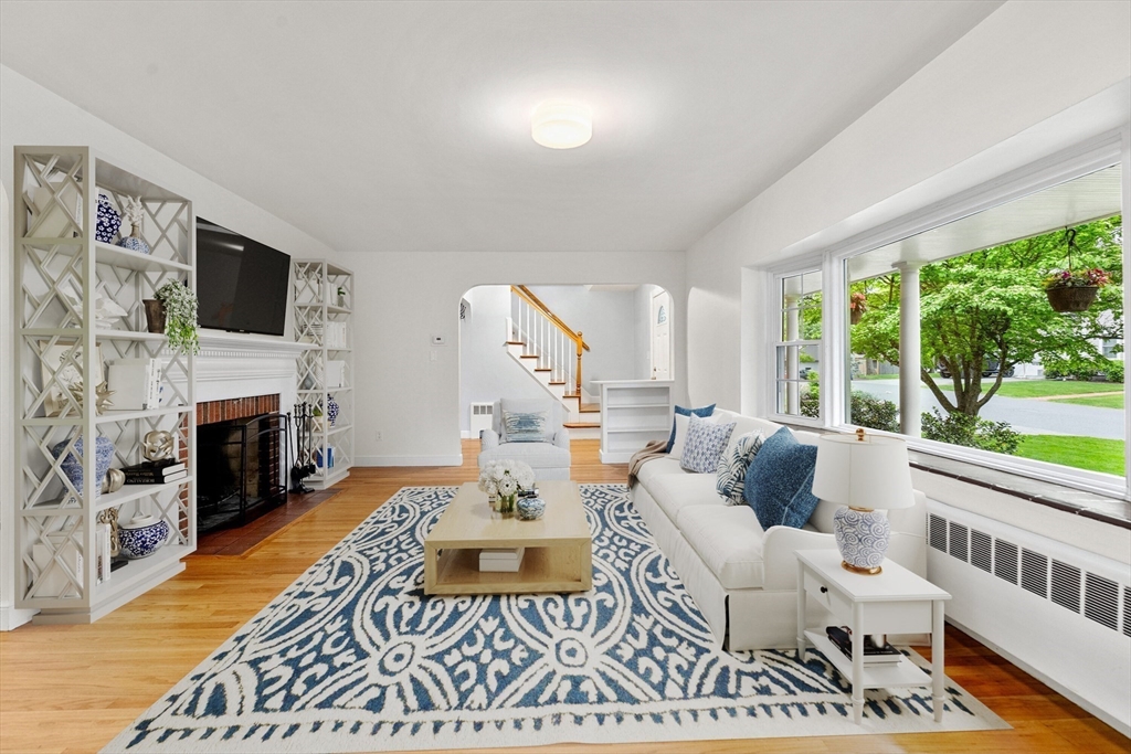 Living room with a fireplace, built-in shelving, white sofas, and large windows overlooking a leafy street.