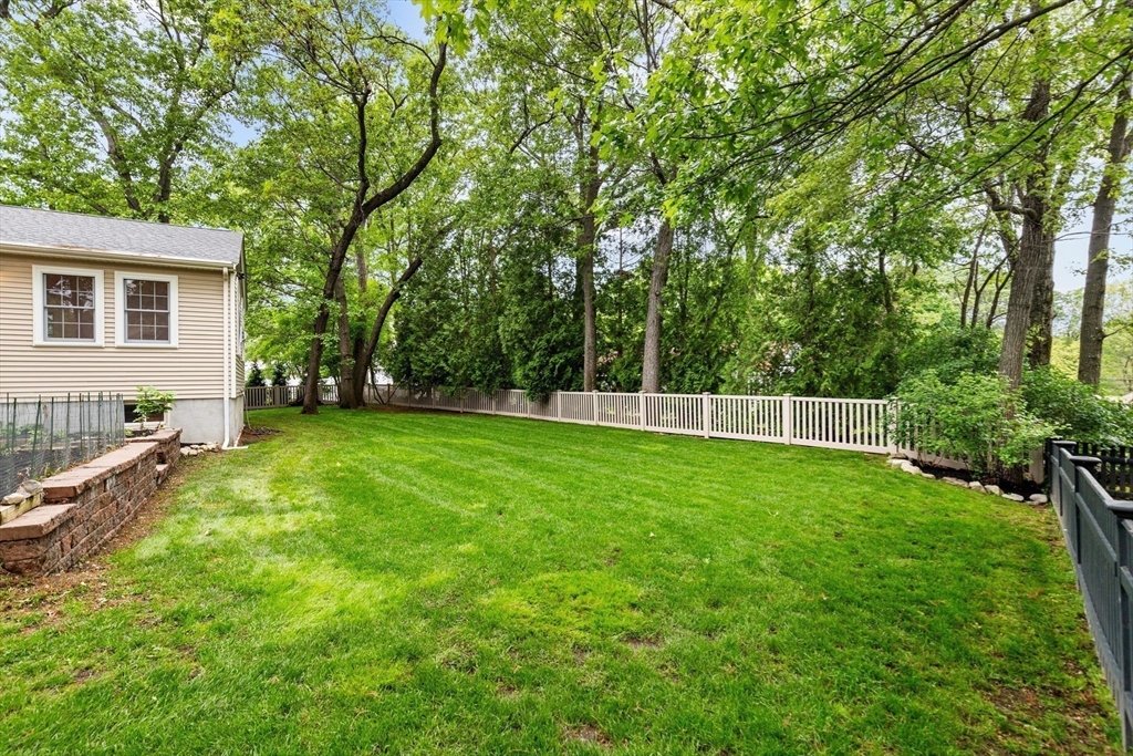 Backyard with a broad lawn, white fence, mature trees, and a stone retaining wall beside the house.
