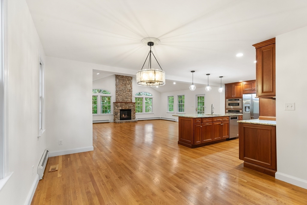 Open-plan kitchen and living area with a stone fireplace, wood floors, and reddish wood cabinetry.