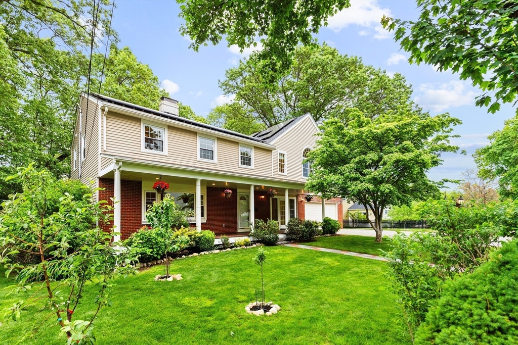 Two-story beige-and-brick home with a covered porch and landscaped lawn framed by leafy trees.