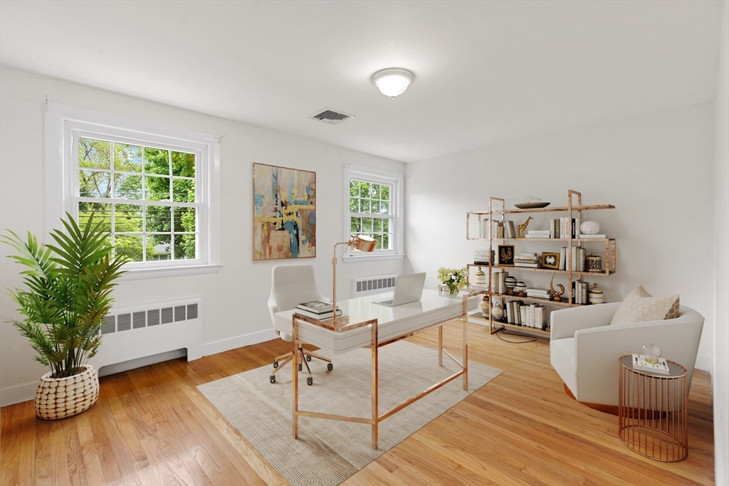 Staged home office with a glass-topped desk, hardwood floors, and leafy views through two windows.