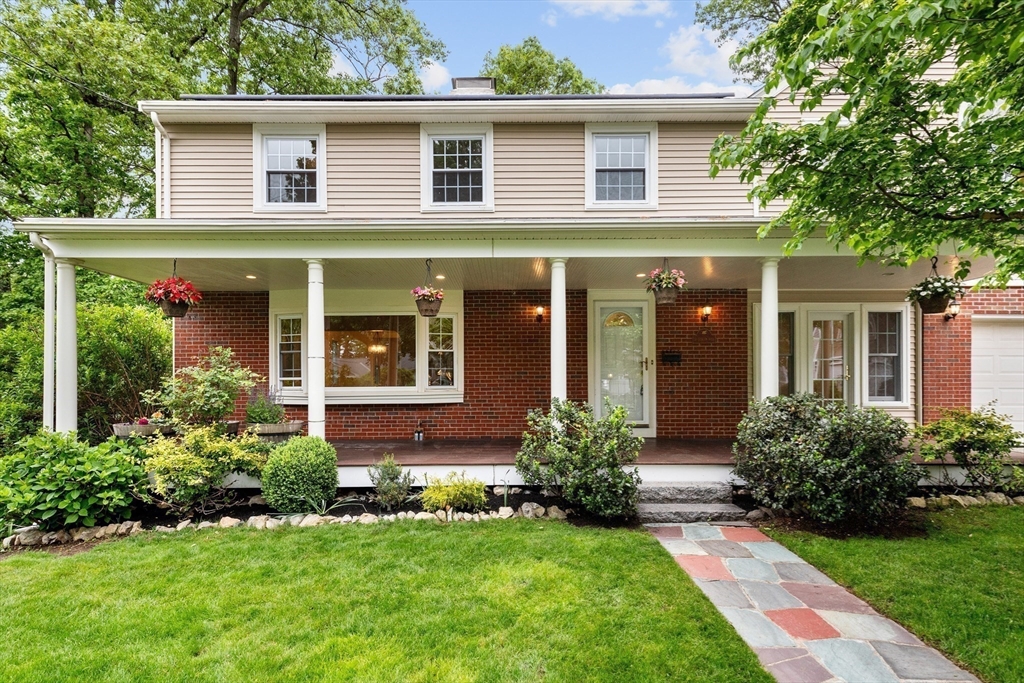 Two-story brick and beige-sided house with a covered porch, hanging flowers, and a stone walkway.