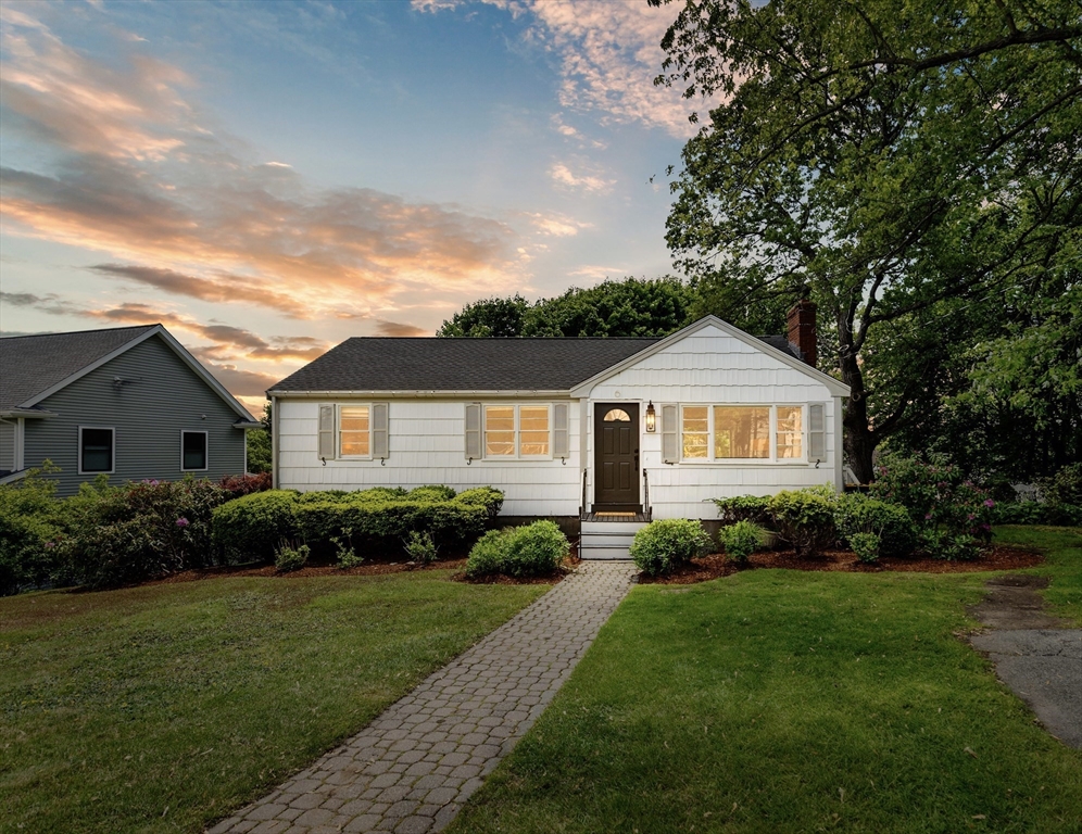 White single-story house with glowing windows, landscaped shrubs, and a stone walkway at sunset.