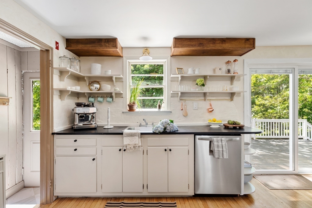 Bright white kitchen with open shelves, wood ceiling beams, and a glass door to a tree-lined deck.