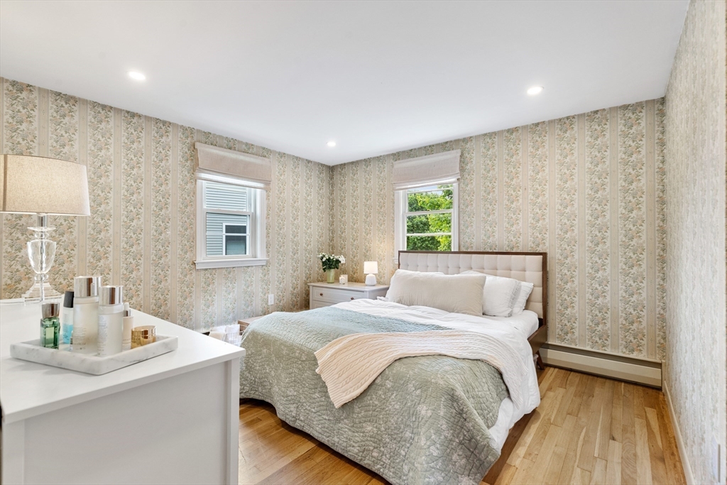 Bedroom with a tufted bed, sage bedding, floral stripe wallpaper, and hardwood floors.