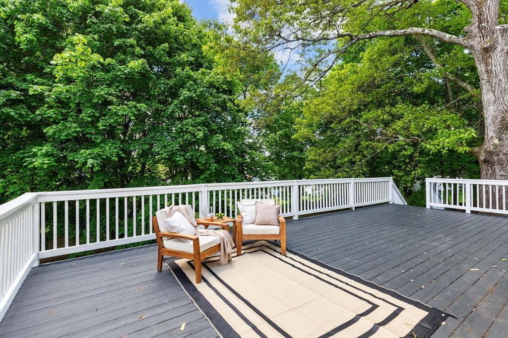 Outdoor deck with white railings, two cushioned chairs, and a rug beneath mature trees.
