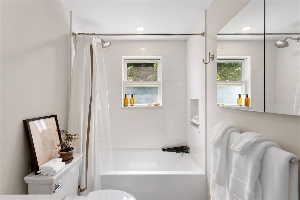 White bathroom with a curtained bathtub, window, mirrored cabinet, and neatly folded towels.