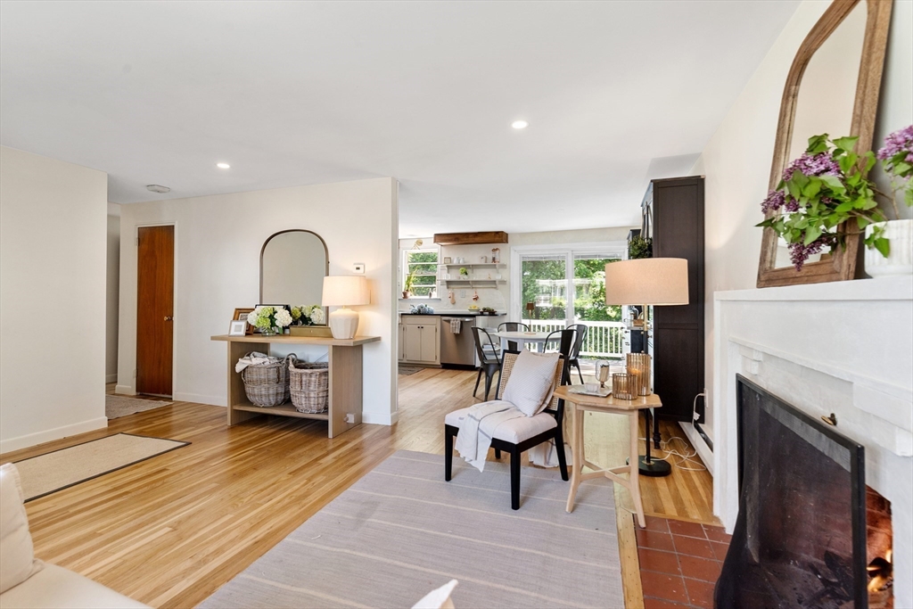 Living room with a white fireplace and hardwood floors, opening to a dining area and kitchen.