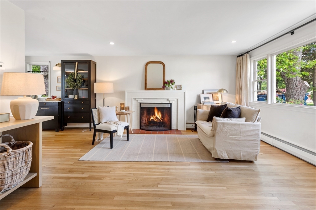 Staged living room with a lit fireplace, hardwood floors, and large windows overlooking greenery.