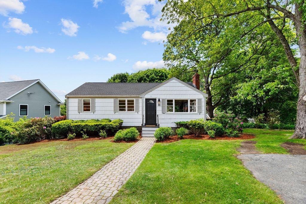 White single-story house with a dark front door, brick walkway, and large leafy tree.