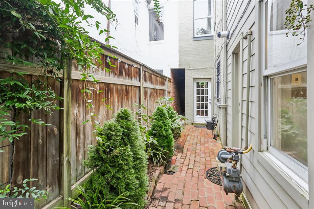 Side passage with a brick walkway, wooden fence, and leafy shrubs beside the house.