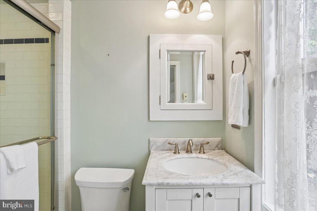 Bathroom with a marble vanity and glass-enclosed tiled shower beside a lace-curtained window.