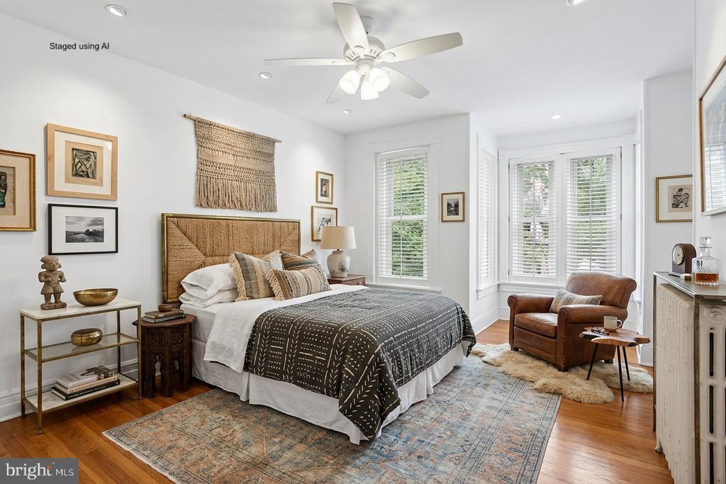 Staged bedroom with a woven headboard, patterned bedding, hardwood floors, and a leather armchair by tall windows.