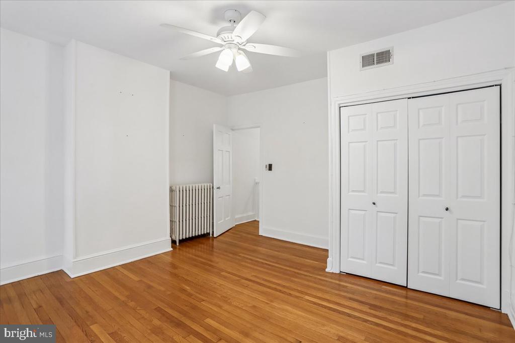Empty room with hardwood floors, a ceiling fan, radiator, and white closet doors.