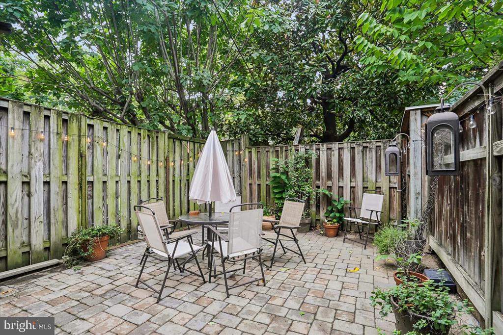 Fenced stone patio with outdoor dining furniture, potted plants, and a closed umbrella beneath leafy trees.