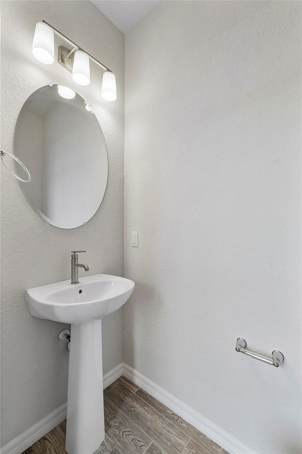 Bathroom with a white pedestal sink, oval mirror, three-light fixture, and wood-look tile floor.