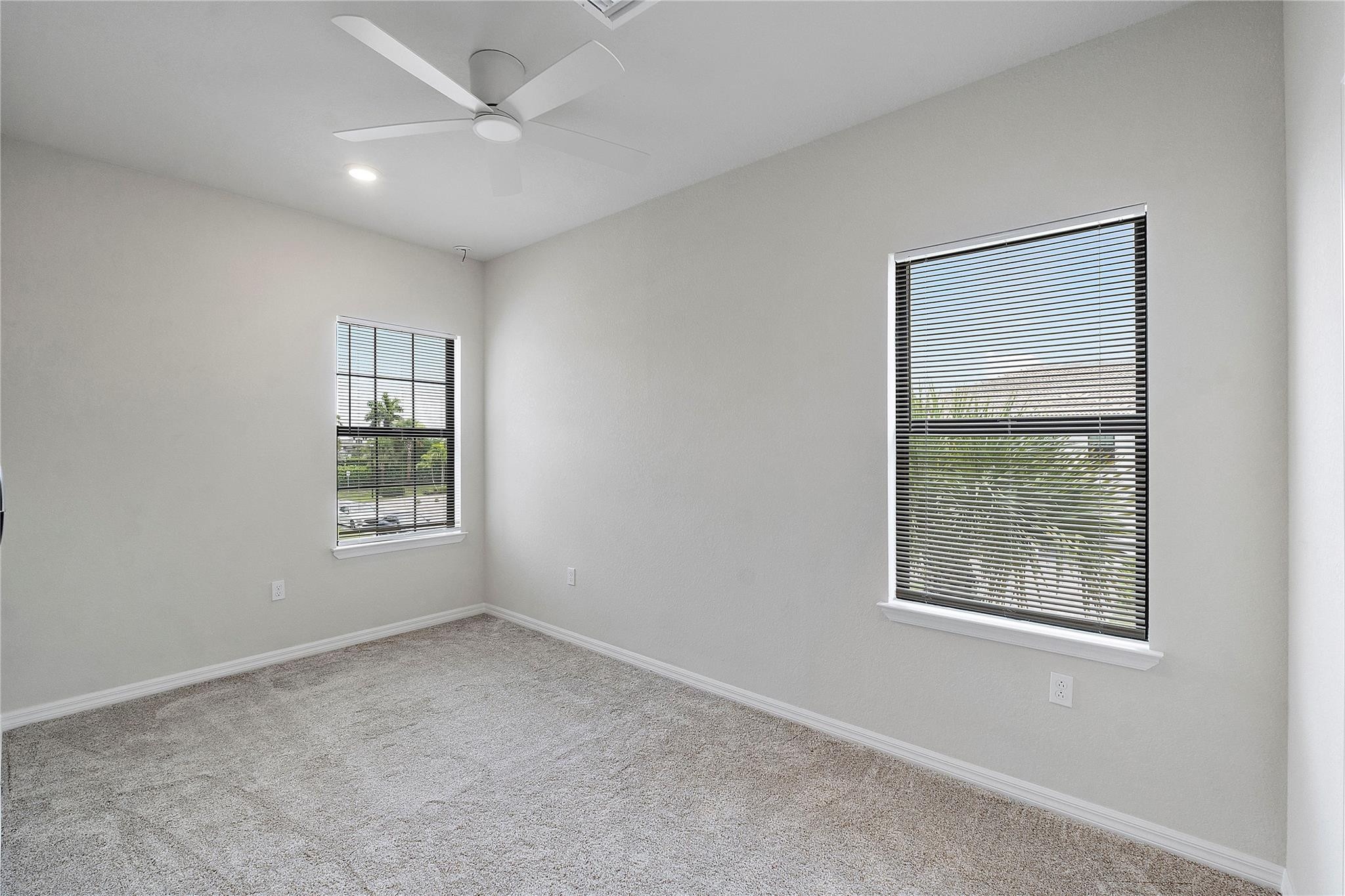 Empty carpeted bedroom with a ceiling fan and two windows with blinds.