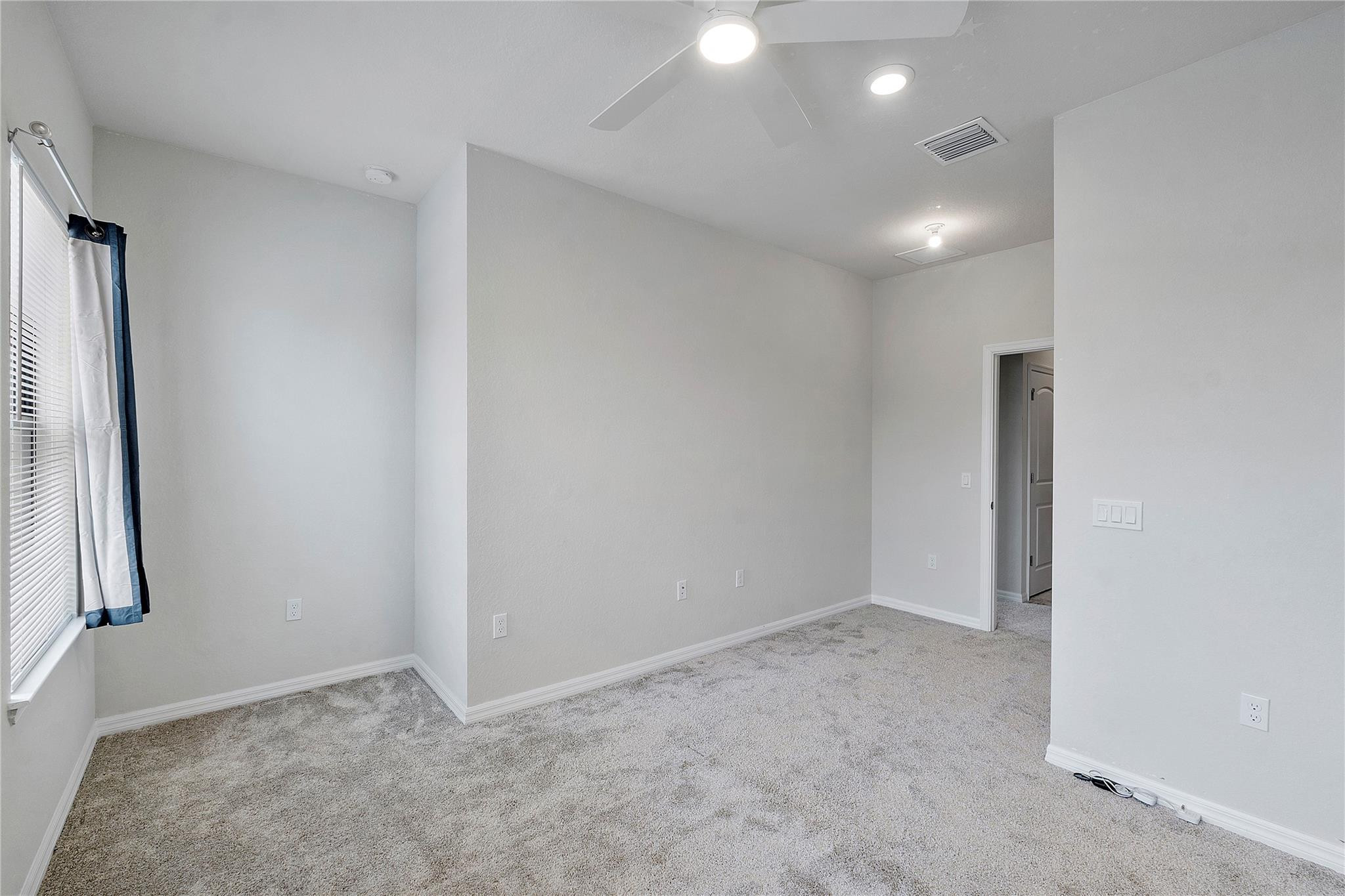 Empty carpeted bedroom with a ceiling fan and a window framed by blue-and-white curtains.