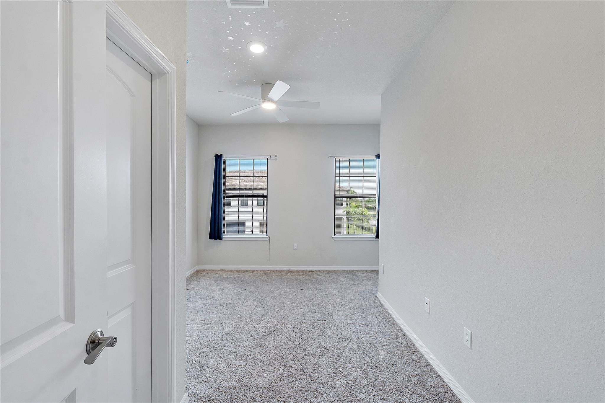 Empty carpeted bedroom with two curtained windows, a ceiling fan, and star decorations on the ceiling.