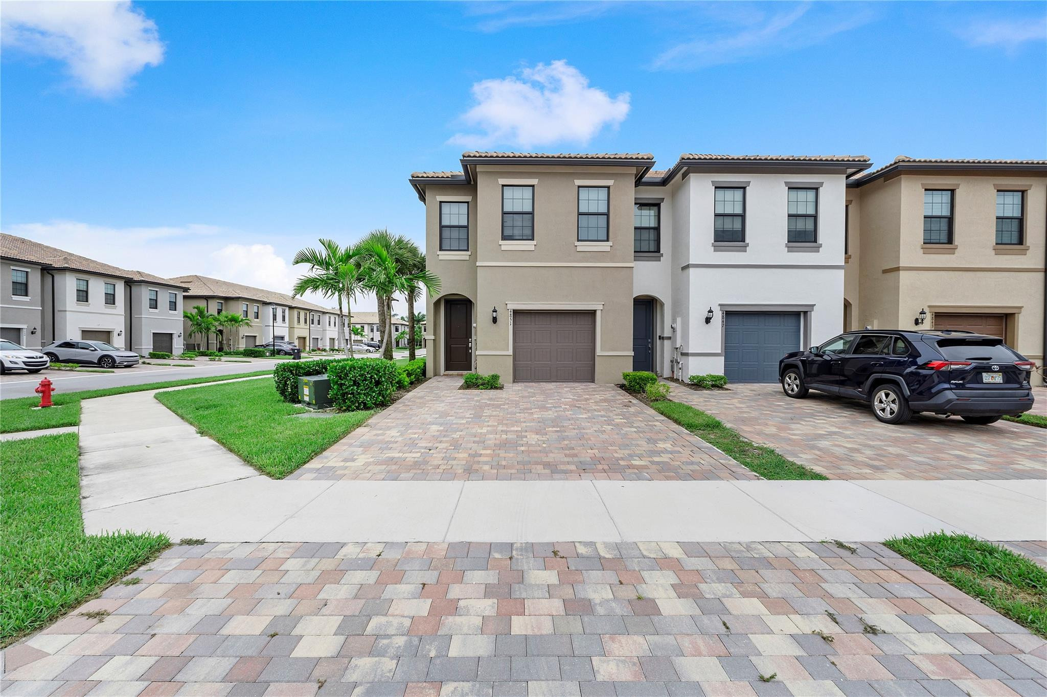 Beige two-story townhouse with a brick-paver driveway, palm trees, and neighboring homes.