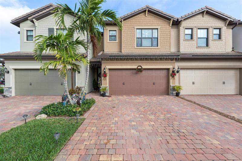Front of a tan two-story attached home with a brick-paver driveway and palm landscaping.