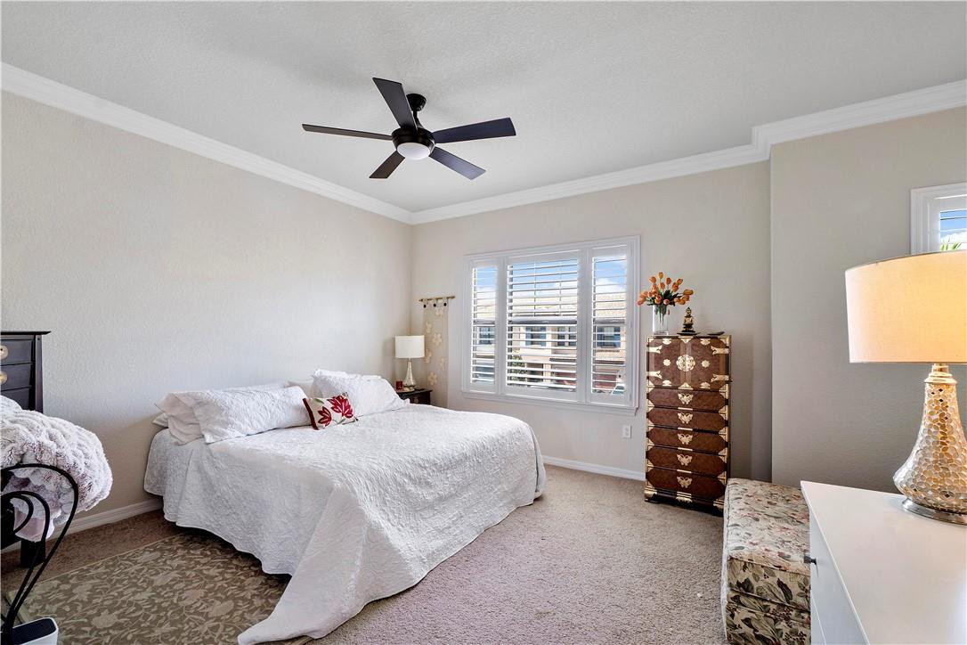 Bedroom with a white bed, dark ceiling fan, plantation shutters, and ornate wooden chest.