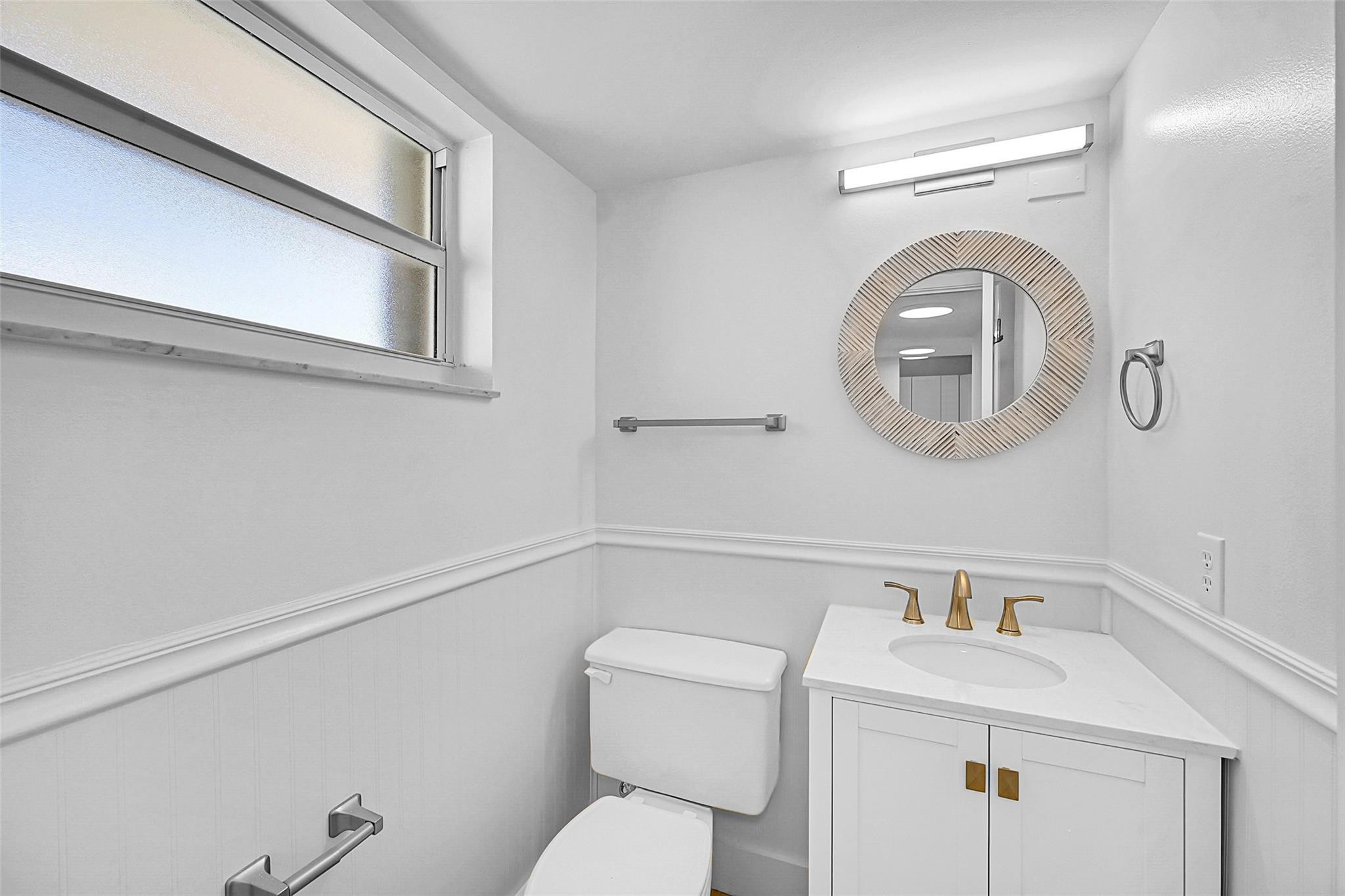 Bathroom with white vanity, gold-tone faucets, round mirror, and frosted-glass window.