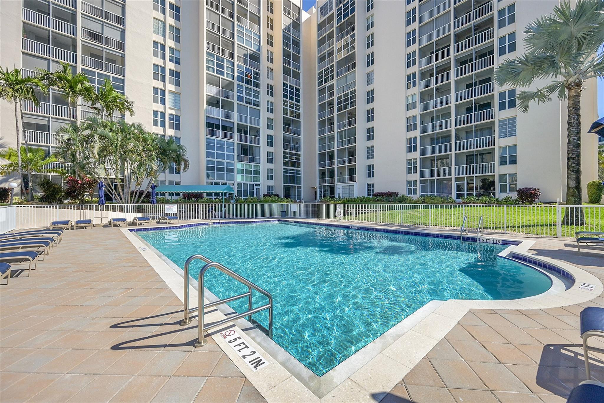 Condominium pool with a tiled deck, lounge chairs, and palm trees.