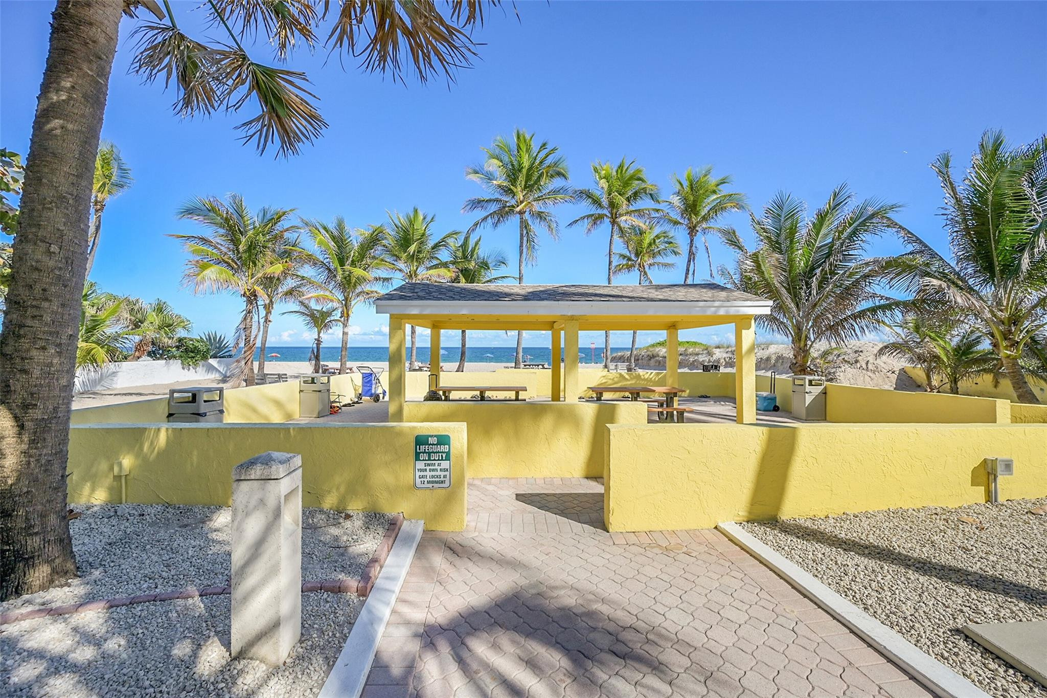 Beachfront pavilion with picnic tables, palms, and a walkway leading to the ocean.
