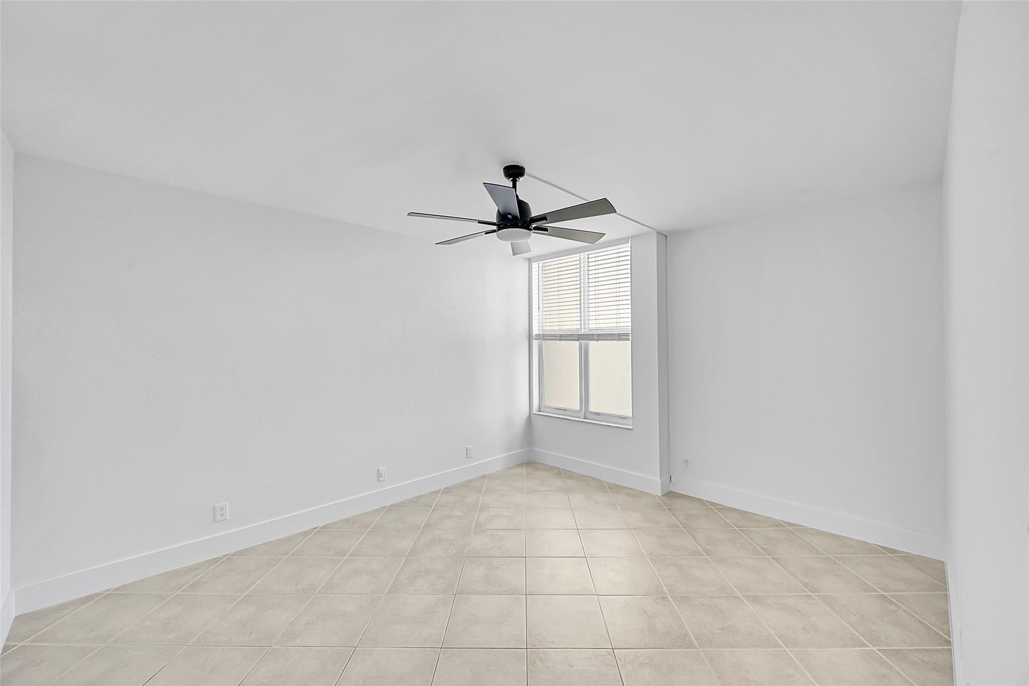 Empty room with beige tile flooring, a dark ceiling fan, and a window with blinds.