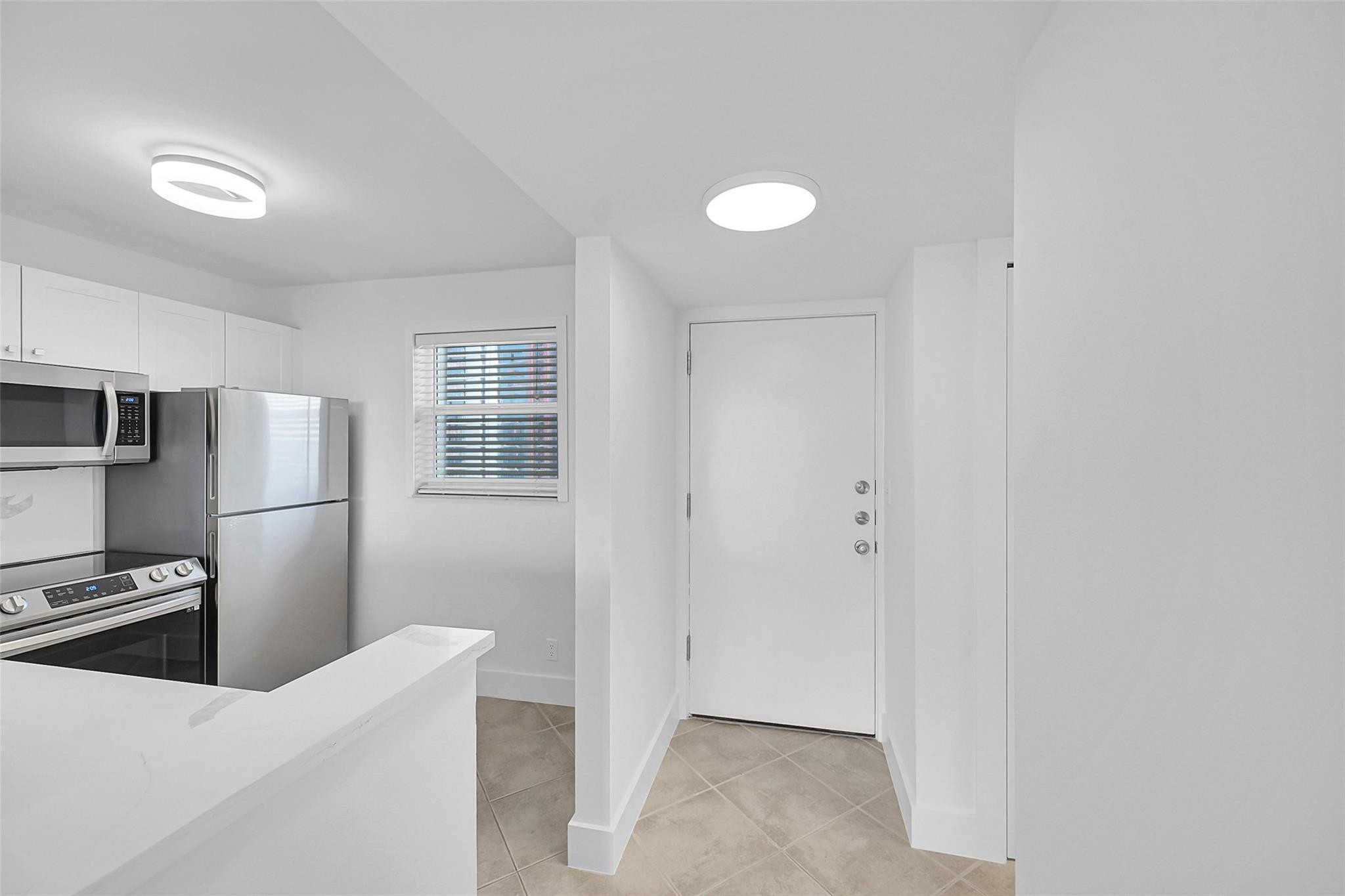 Kitchen and entry with stainless steel appliances, white peninsula countertop, and beige tile flooring.
